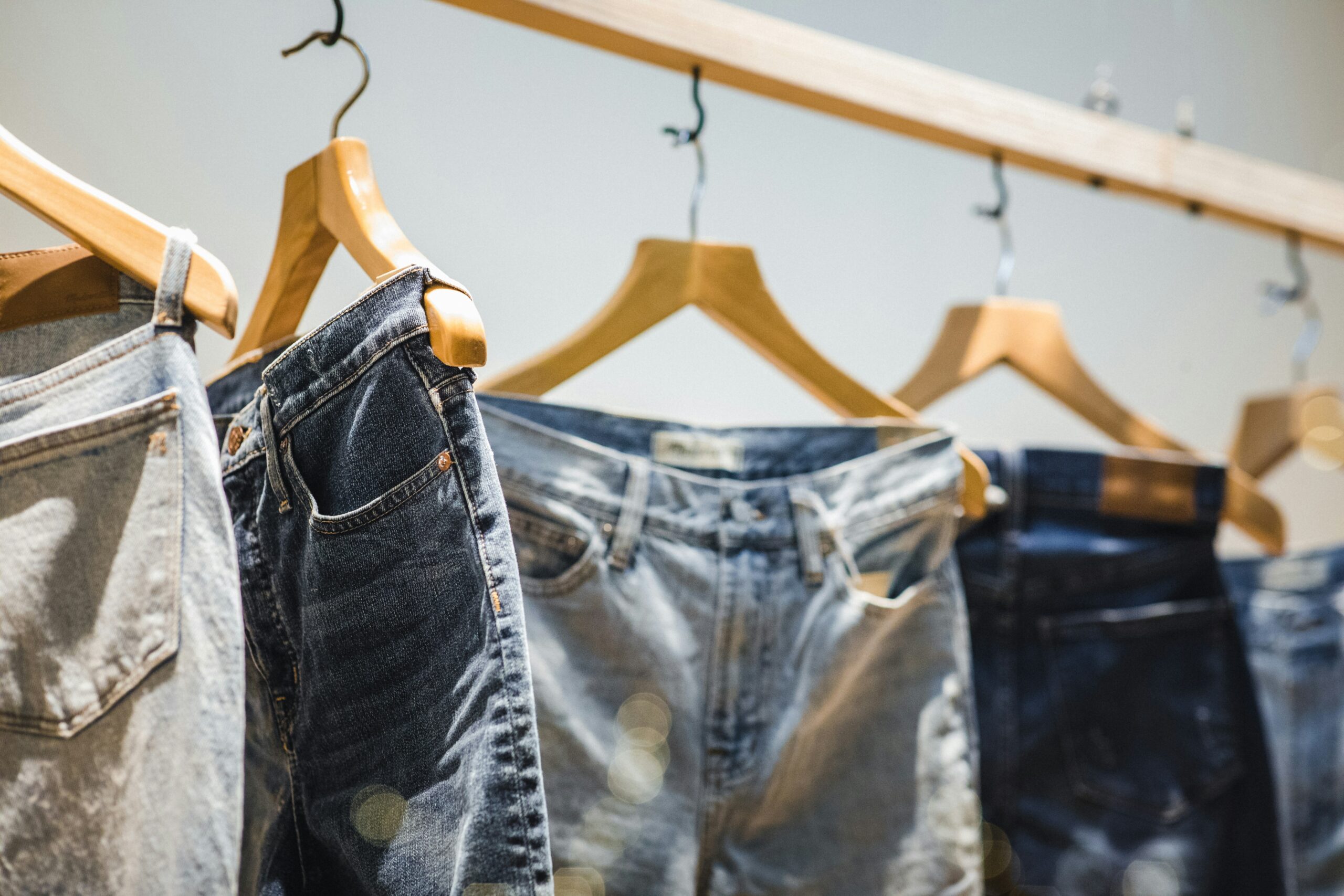 A perspective shot of several pairs of jeans in various blue washes hanging from a wooden clothing rack. The jeans are suspended by wooden hangers, with the pair in the foreground showing a dark indigo wash and clear pocket detailing.